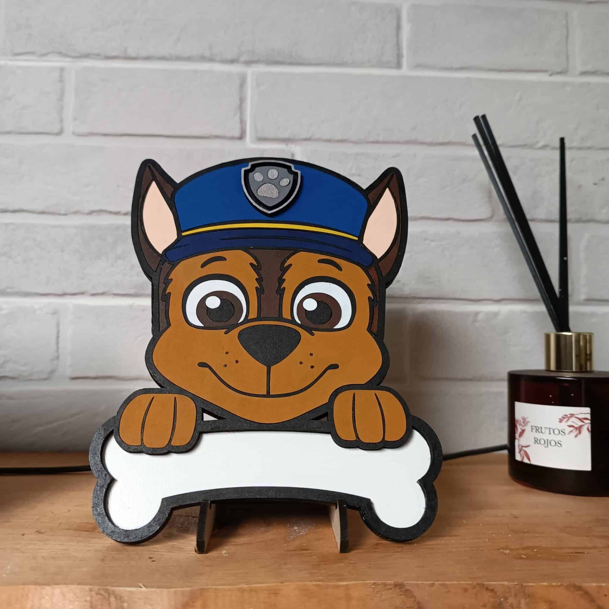 cartel del dibujo animado Chase de la patrulla canina. Es un perro marrón con una gorra azul con su escudo de una huella plateada. Chase está sujetando un hueso donde alli va pegado el nombre del niño o niña.